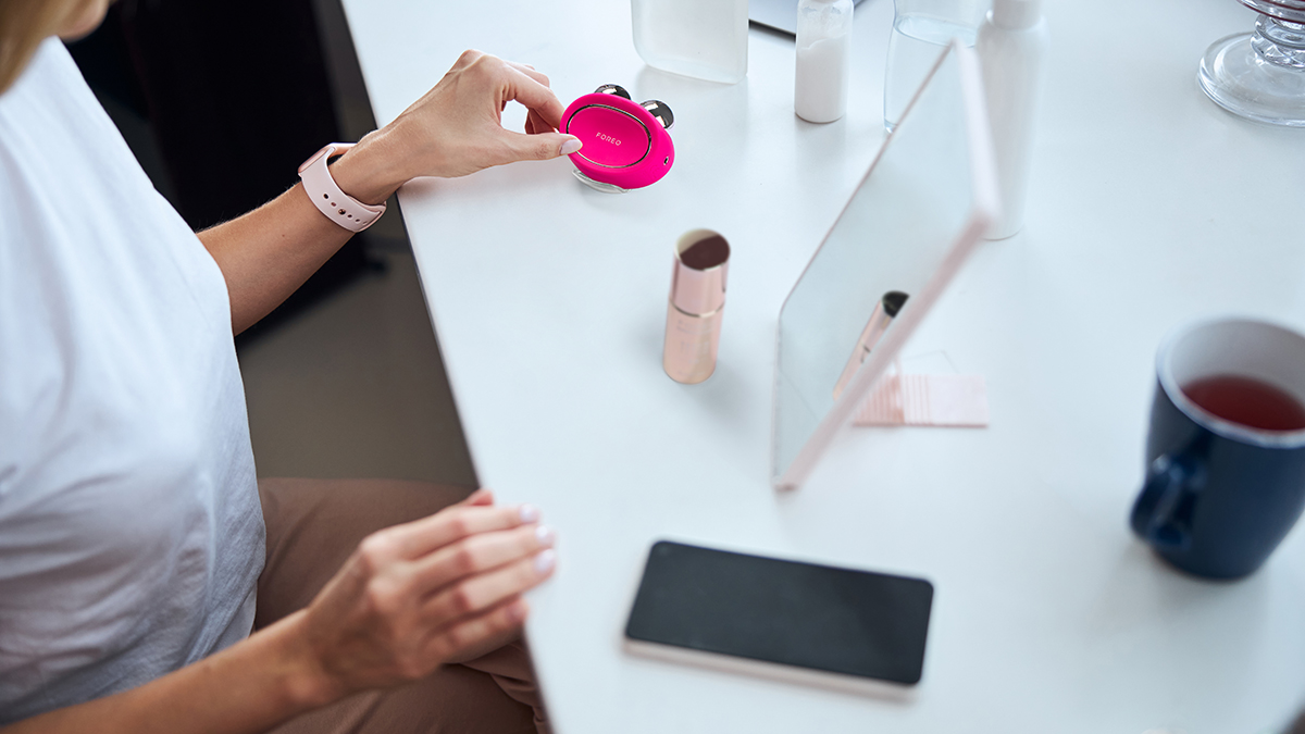 Photo with FOREO BEAR 2, FOREO SUPERCHARGED 2.0 SERUM, cell phone, a mirror, and hands on the table