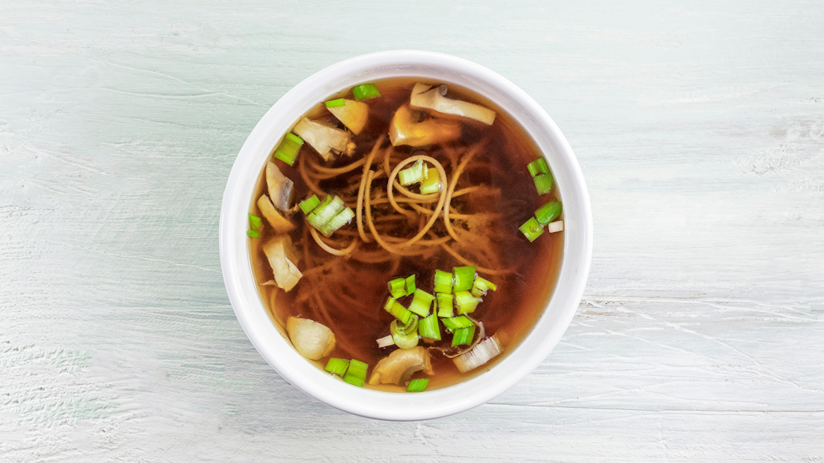 A bowl of miso soup with scallions, mushrooms, and soba noodles on the table