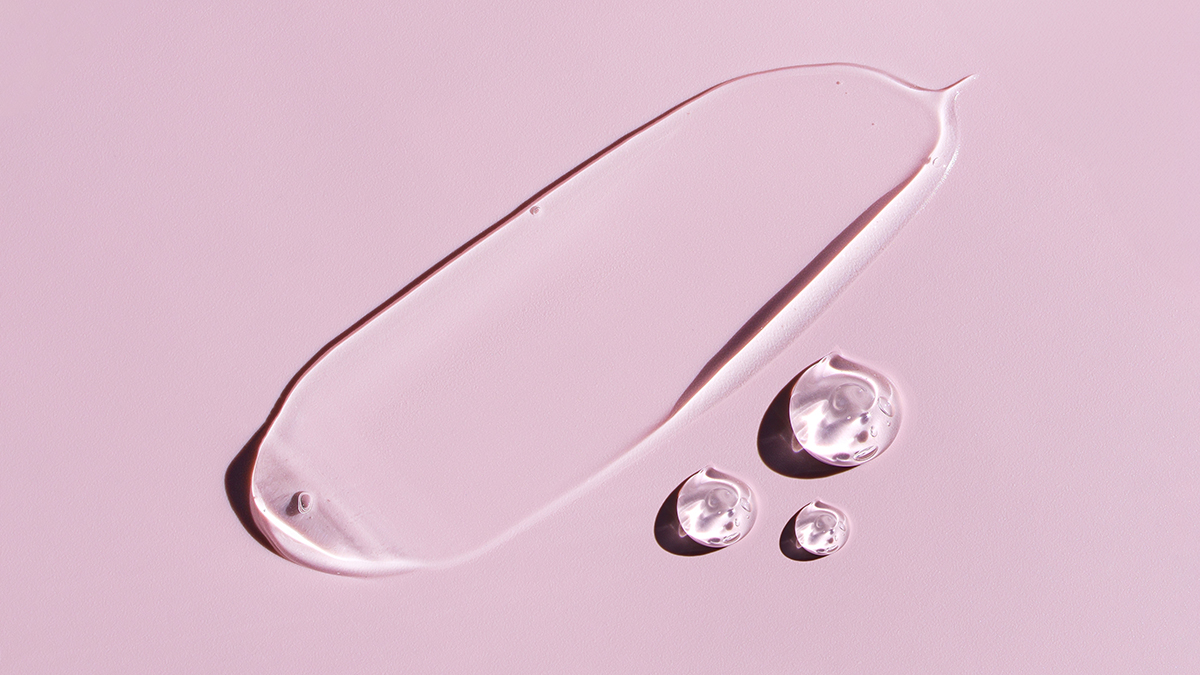 Transparent cosmetic gel or serum smeared over a pink surface alongside three gel drops