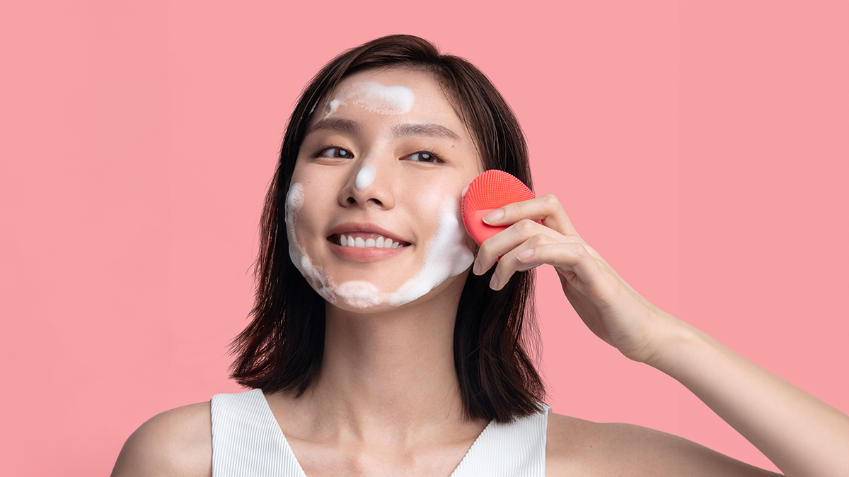 A model cleansing her face with microfoam cleanser and LUNA 4 mini facial cleanser on a peach-colored surface