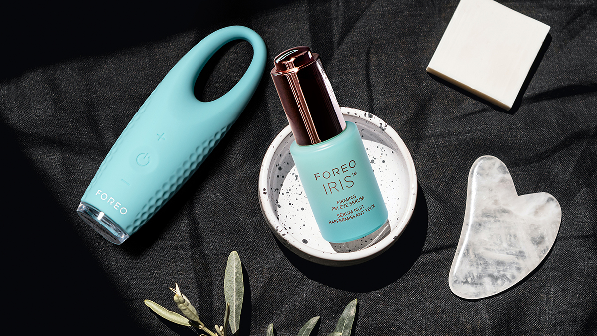 IRIS Firming PM Eye Serum on a table with FOREO IRIS eye massager, olive branch, a heart-shaped object and a square object on black surface