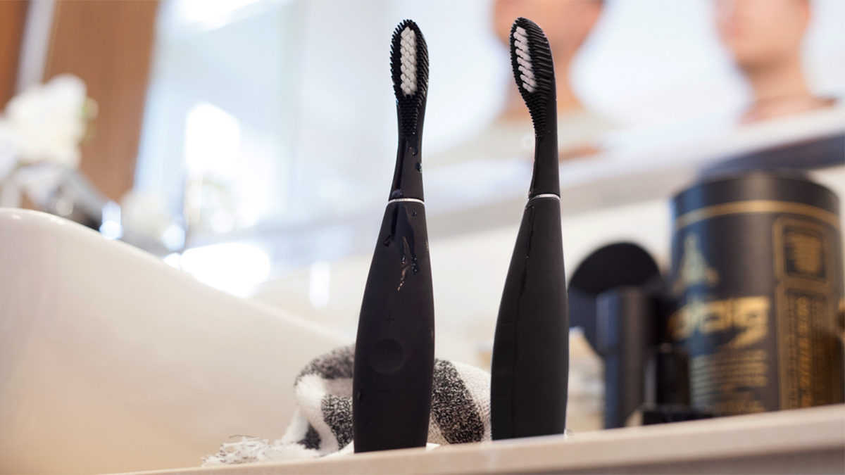Two black FOREO ISSA electronic toothbrushes on a bathroom counter, with two blurry men figures in the back