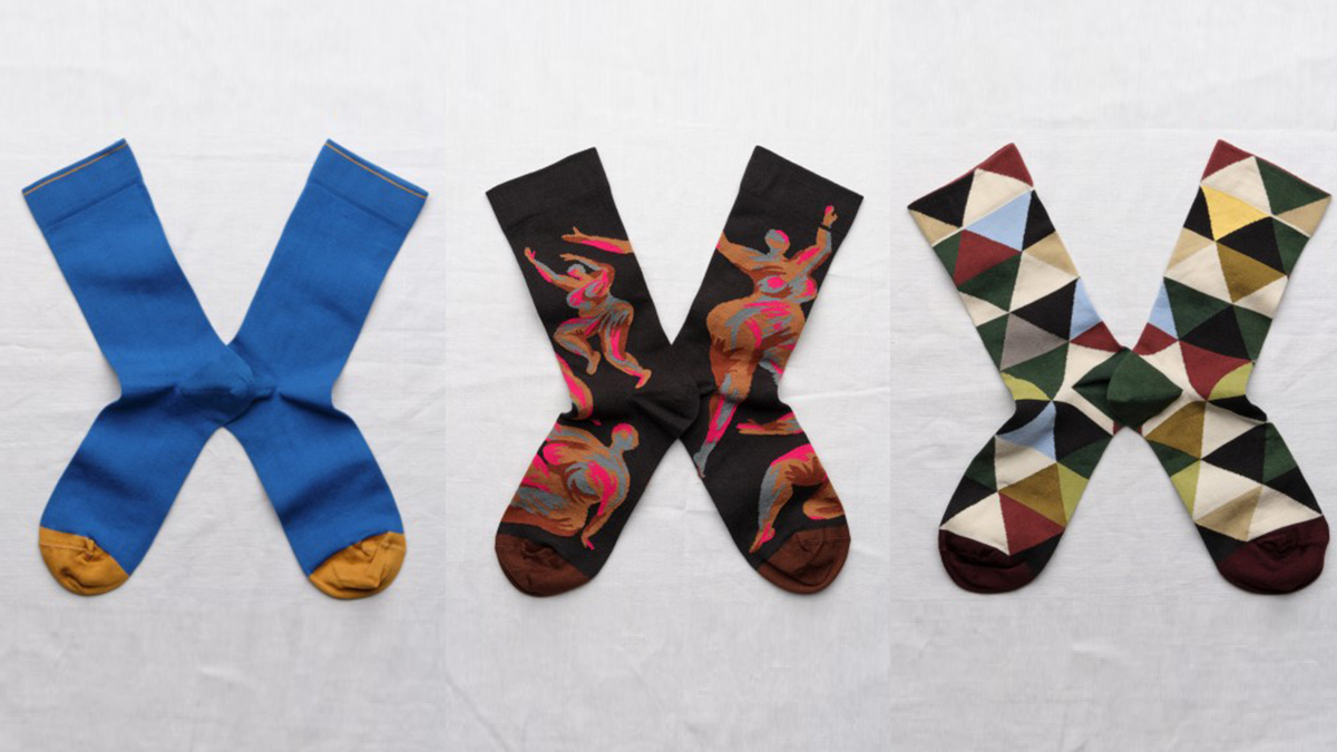 Three pairs of colorful socks on neutral surface: blue with yellow tips, dark with colorful nude women motif, and geometric in green, blue, beige, red, and black