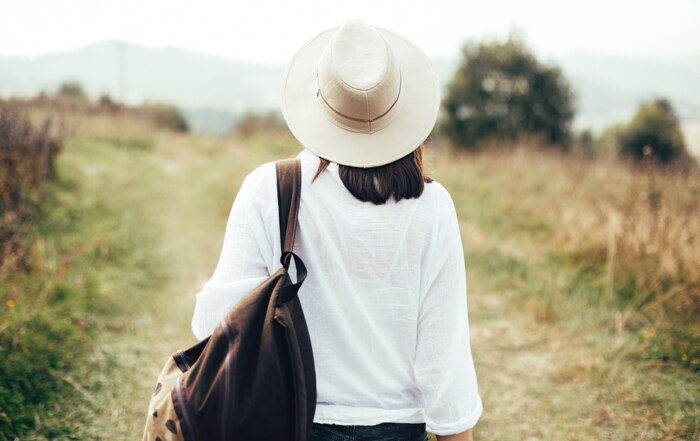 A woman with a hat, white shirt, and a backpack, looking at a countryside road covered in grass