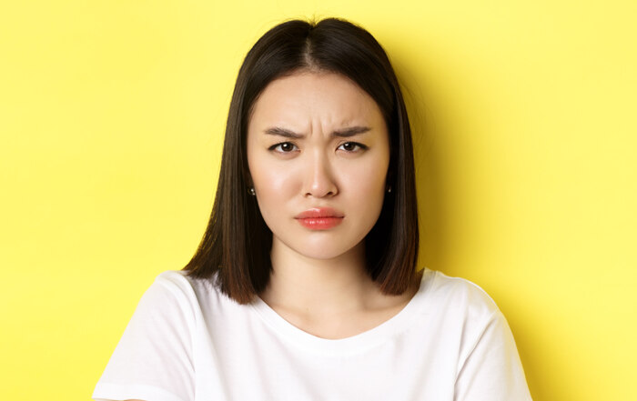 Close up of young Asian woman looking angry and frowning at camera, standing in white t-shirt on yellow background