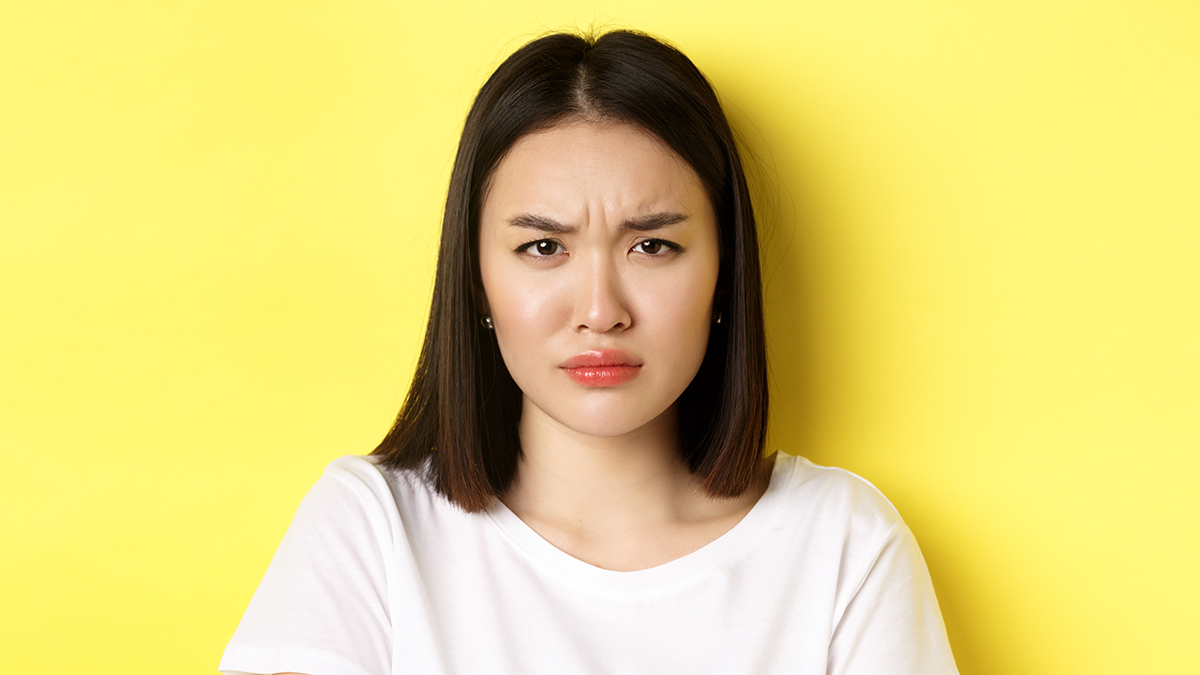 Close up of young Asian woman looking angry and frowning at camera, standing in white t-shirt on yellow background